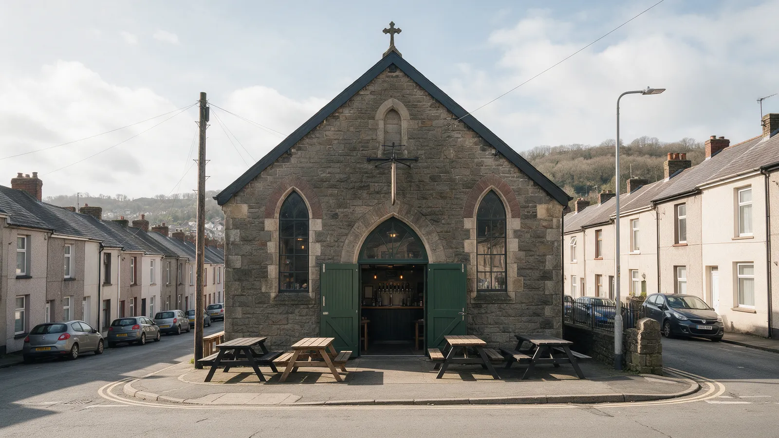 The exterior of a small microbrewery taproom in a converted Victorian stone chapel on a terraced street in Pontypridd, dark green doors open, picnic benches outside, hills behind