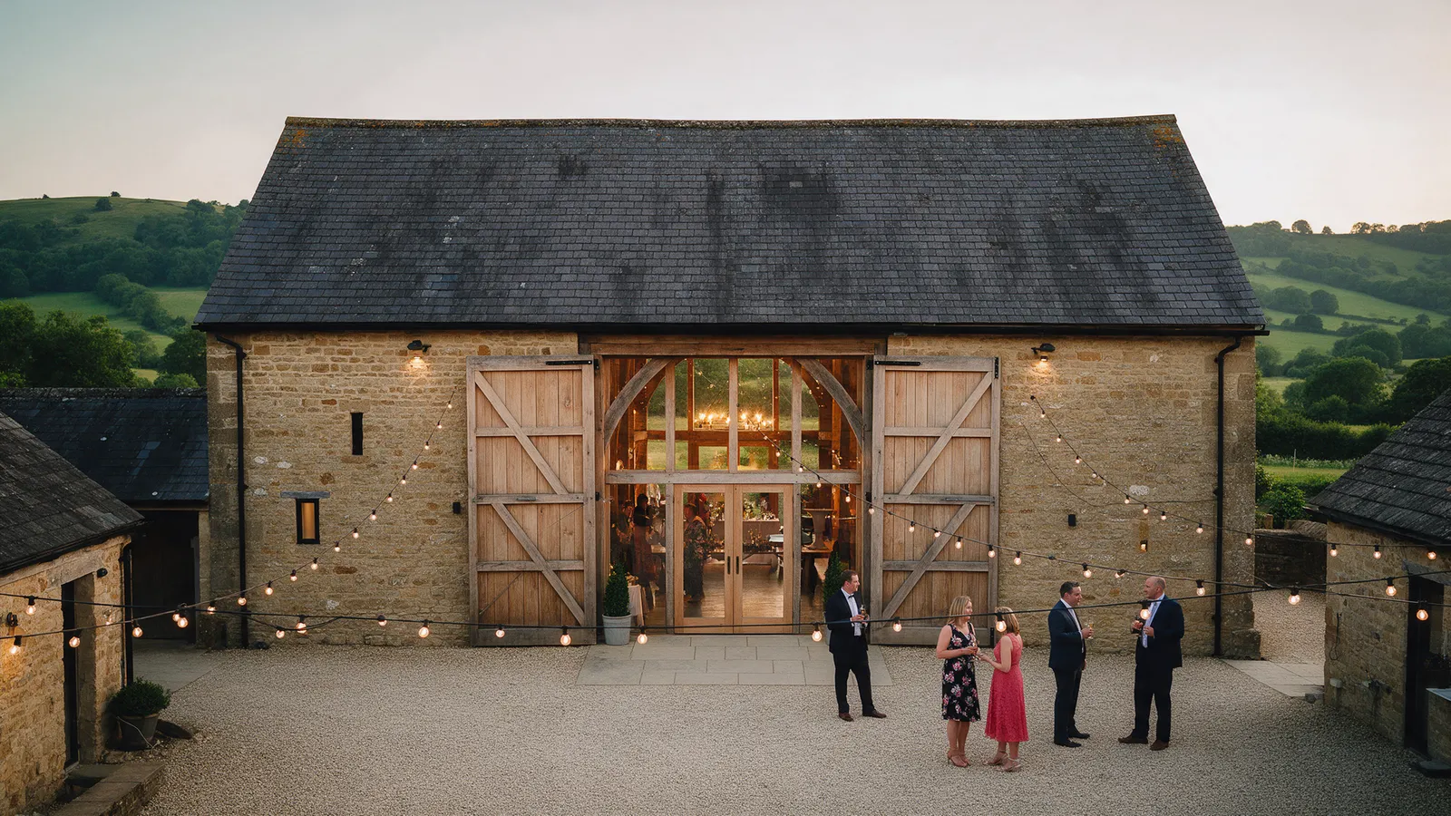 Ty Mawr Barn at dusk: honey-coloured stone, the oak doors open and glowing, festoon lights across the courtyard and the Usk valley behind