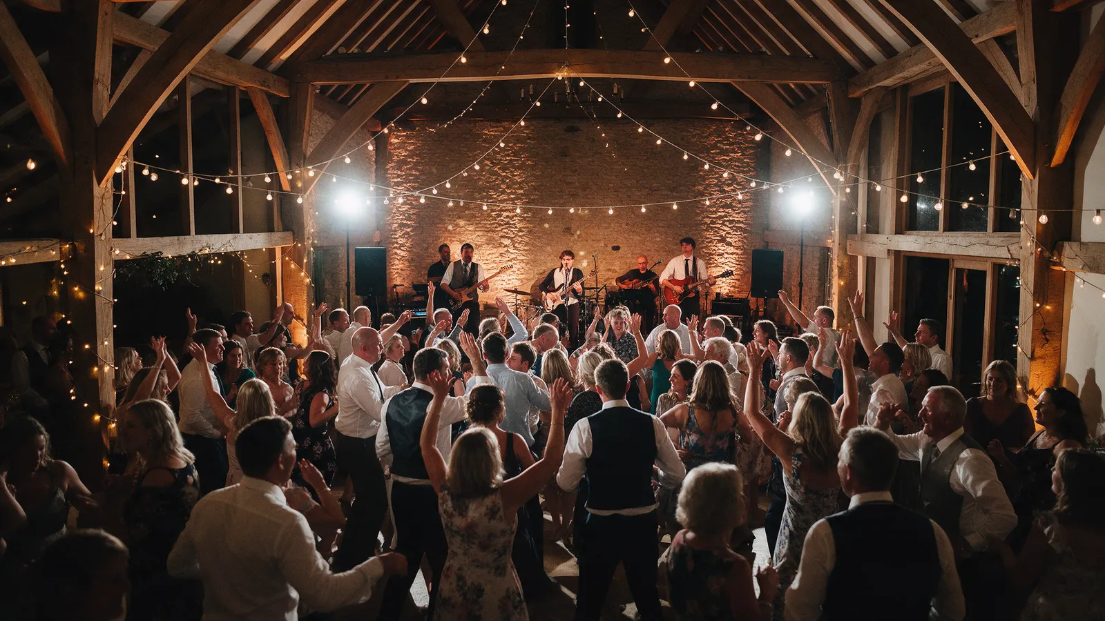 A packed dance floor in the Barn at night with a live band on the stage under festoon lights